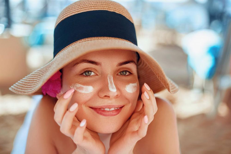A woman wearing a sun hat applies sunscreen to her face